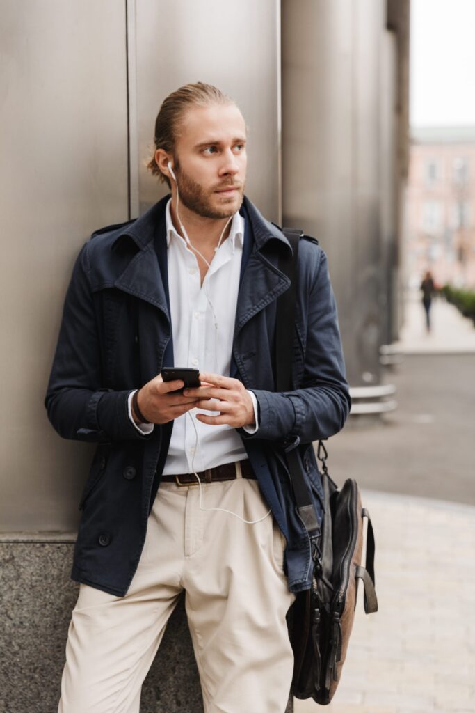 Homme en caban croisé bleu marine et pantalon beige, adossé à un immeuble